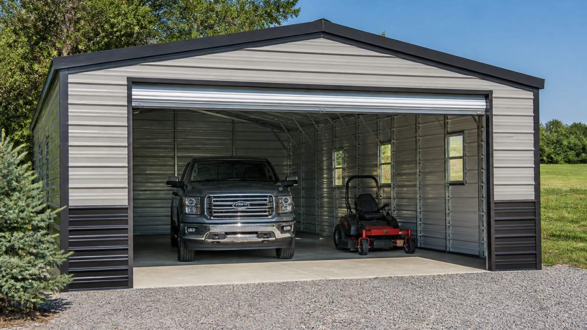 A newly assembled steel garage kit on a rural residential property, with a pickup truck and riding mower parked inside and an open roll-up door showing the clear span interior