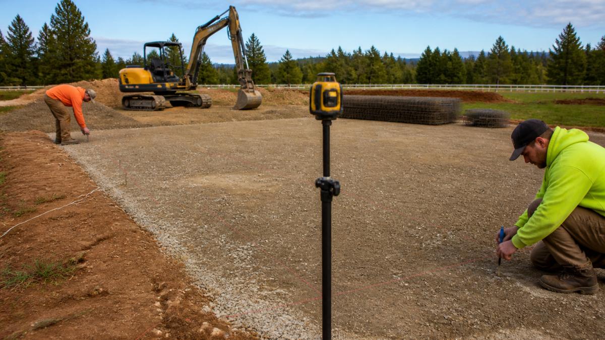 A construction crew using a laser level and string lines to prepare a gravel pad for a steel garage foundation, with a small excavator and steel rebar bundles visible in the background on a rural property