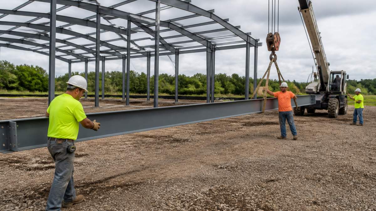 A crew of workers in safety gear guiding a steel beam into place with a small crane on a rural building site, with a partially assembled steel frame in the background