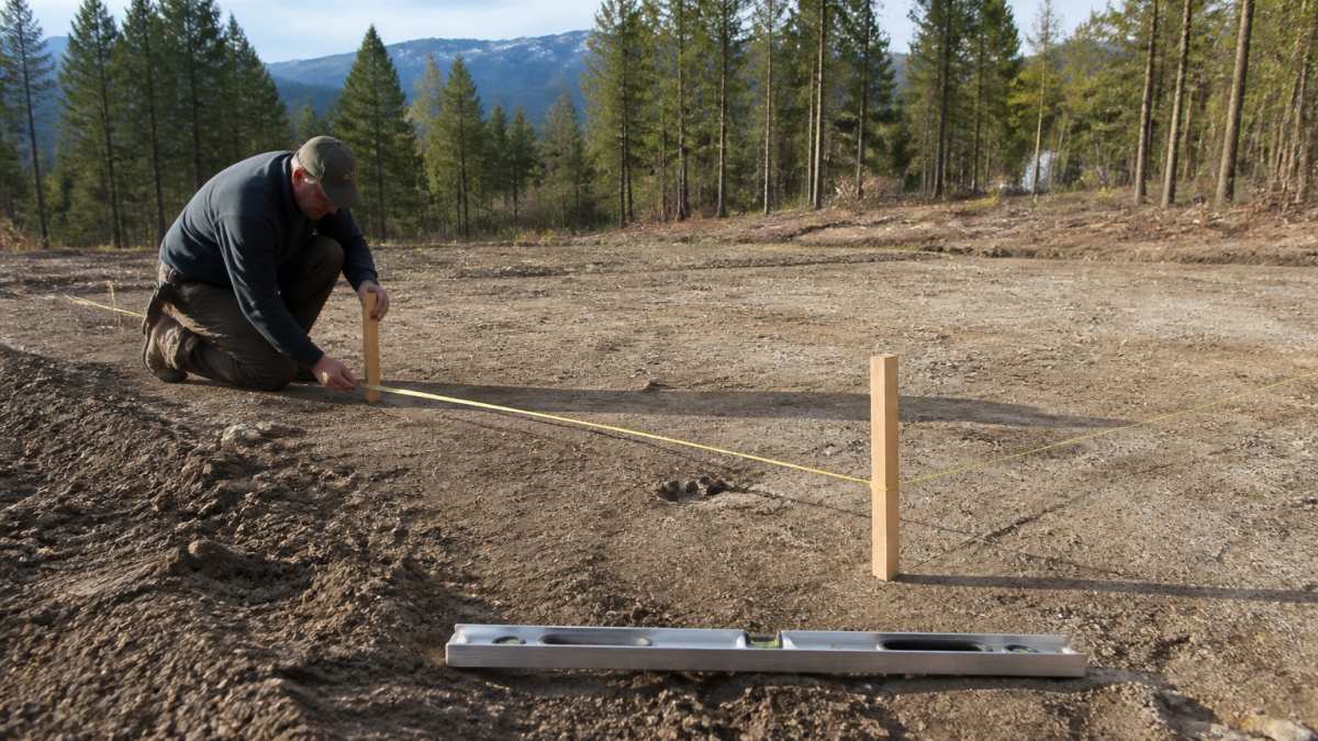 Homeowner using a tape measure and stakes to layout a steel garage foundation on a cleared rural lot